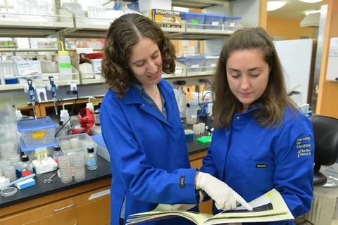 A trainee and a mentor review notes together, both in blue lab coats